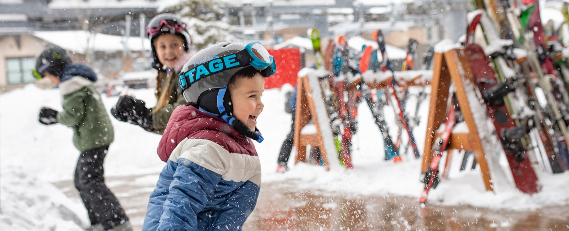 Three young kids wearing ski and winter gear smile and play in the falling snow.