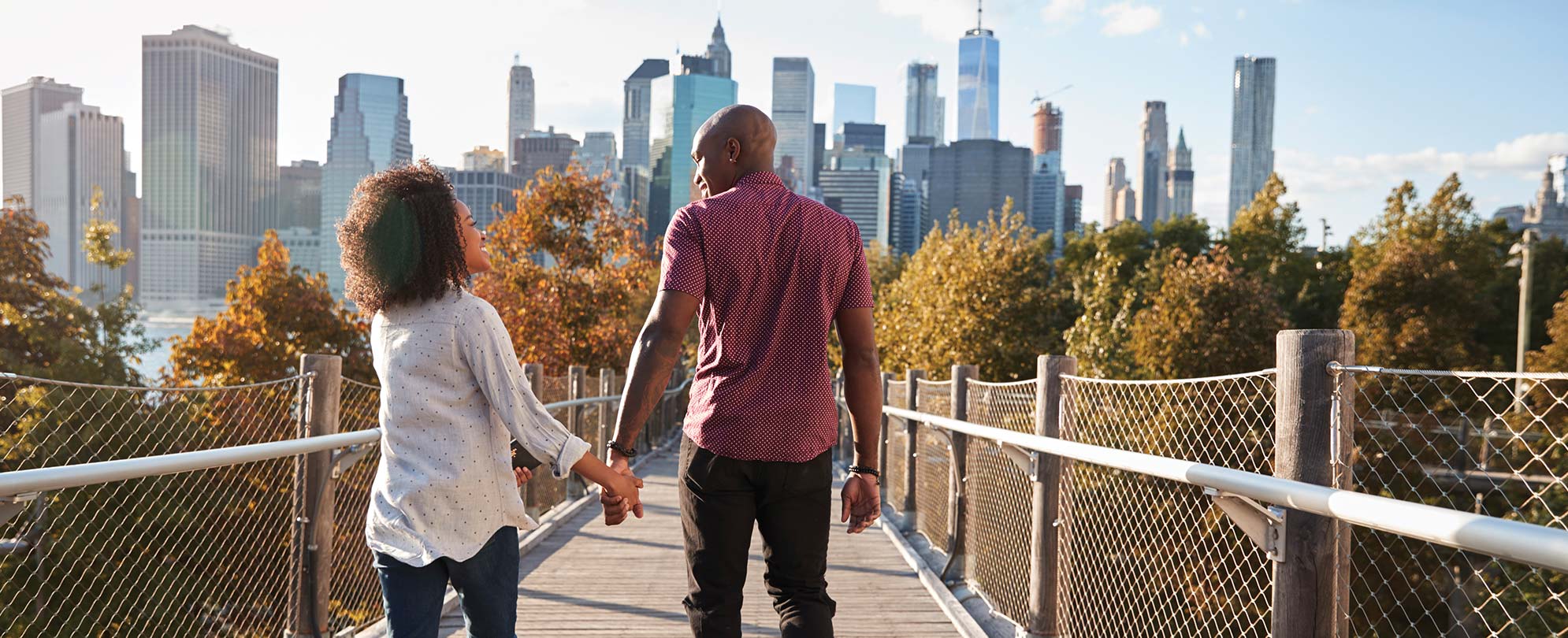A man and a woman hold hands and walk along a boardwalk toward a city skyline