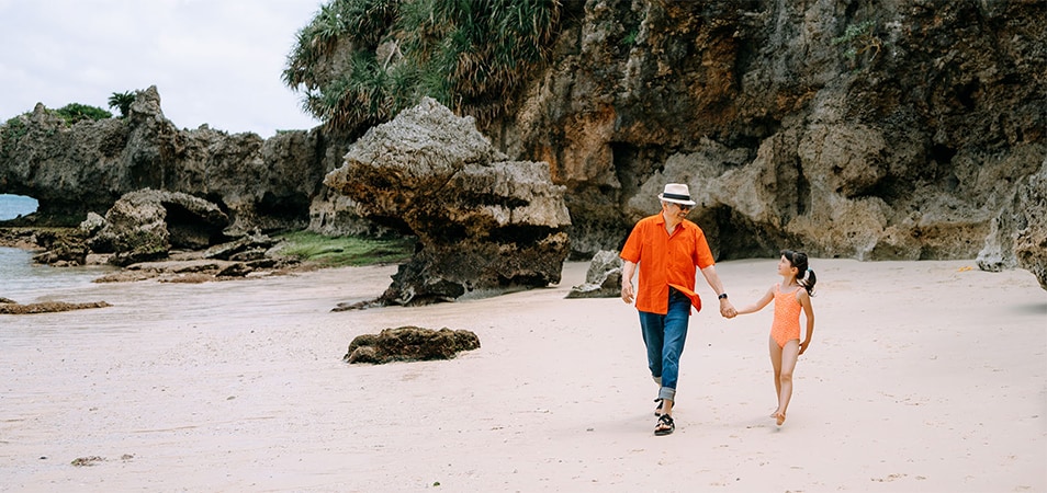 A dad and his young daughter walk holding hands on a beach, rocky cliffs in the background. 