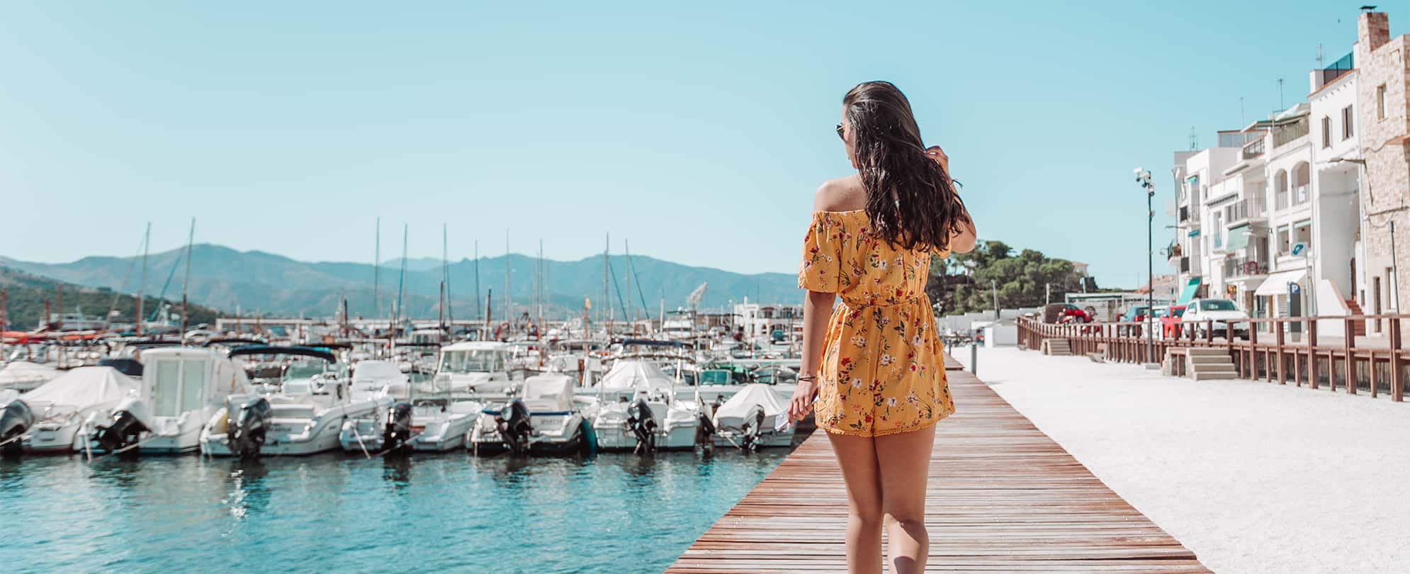 A woman in a yellow romper walks along a marina dock on a summer day