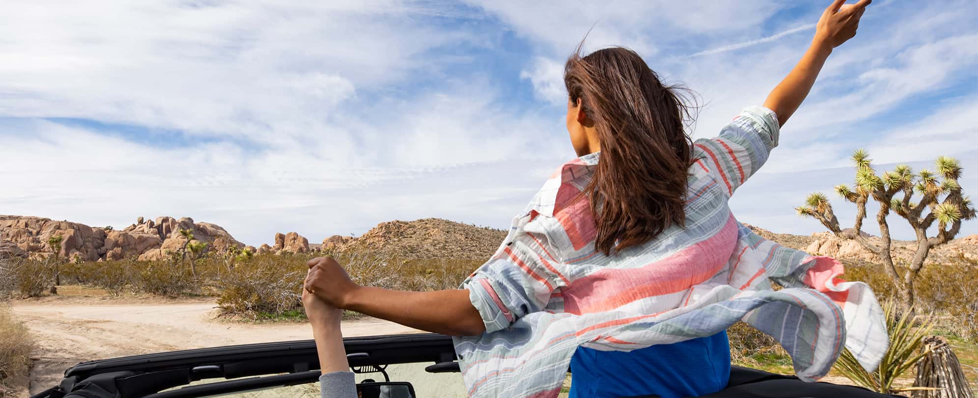 A woman raises her hands out of the top of a open-top convertible while on a road trip through the desert