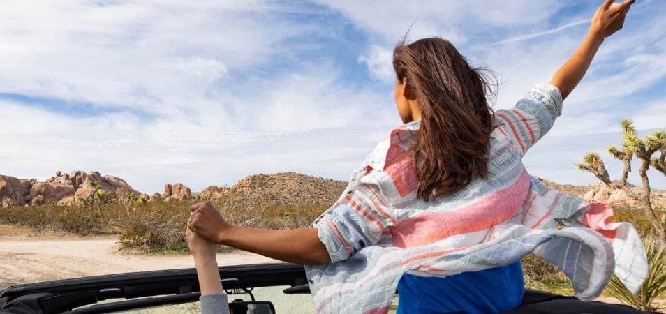 A woman raises her hands out of the top of a open-top convertible while on a road trip through the desert