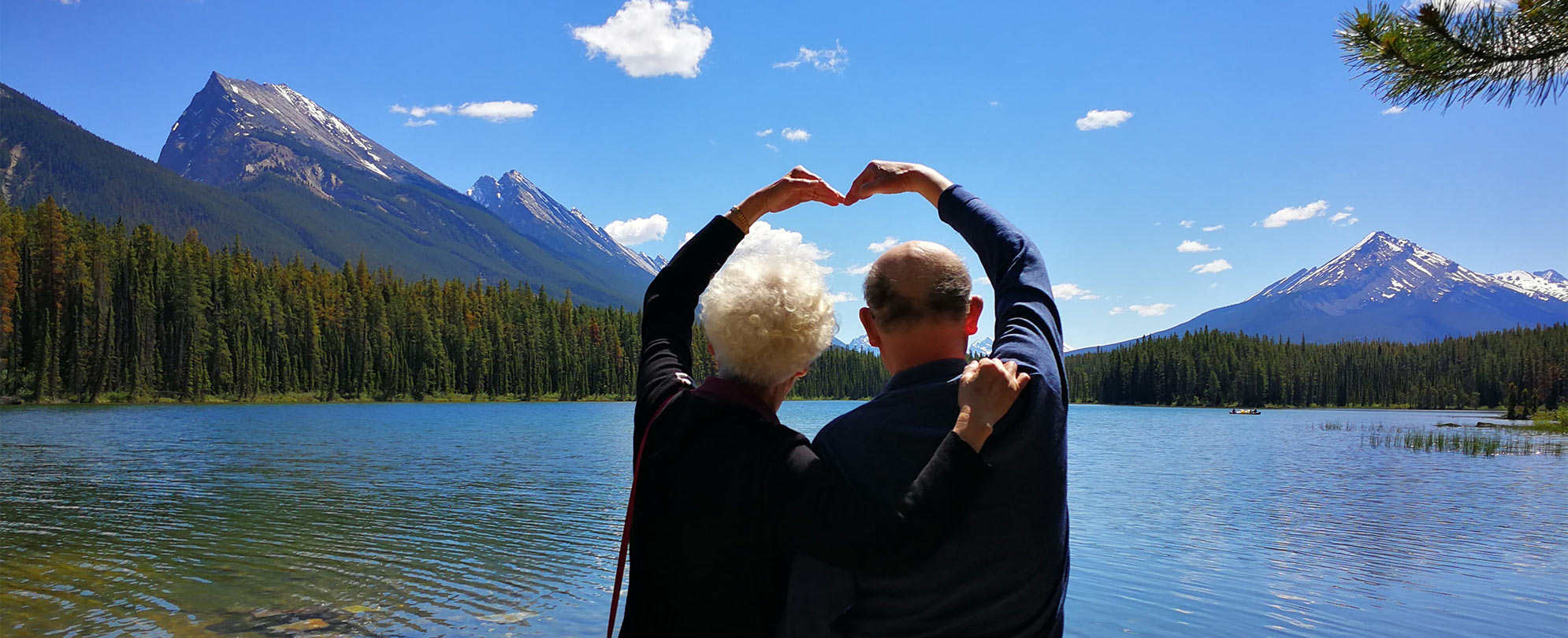 Two Club Wyndham owners pose with their hands forming a heart overlooking mountains and a lake at Banff National Park in Alberta, Canada