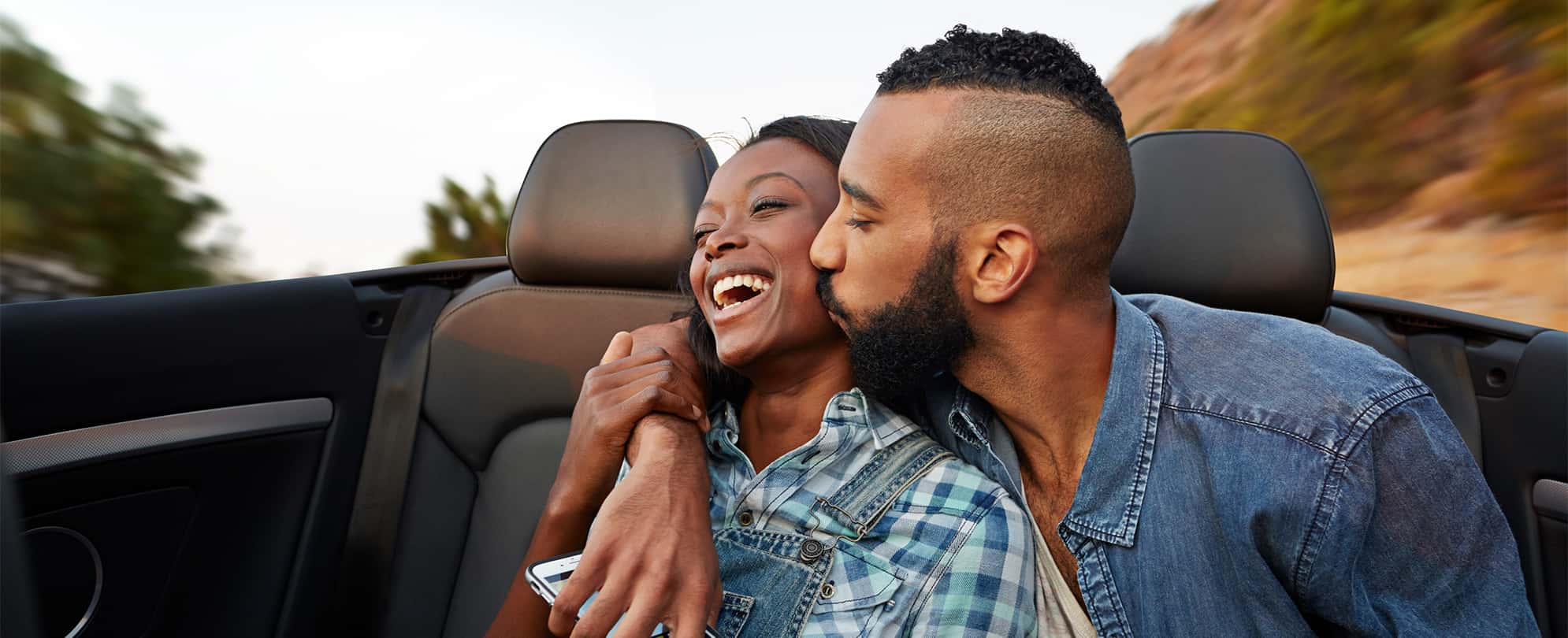 A man kisses a woman on the cheek as they drive their convertible on a road trip vacation