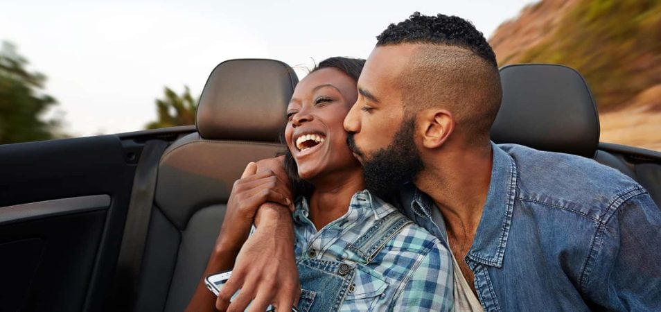 A man kisses a woman on the cheek as they drive their convertible on a road trip vacation