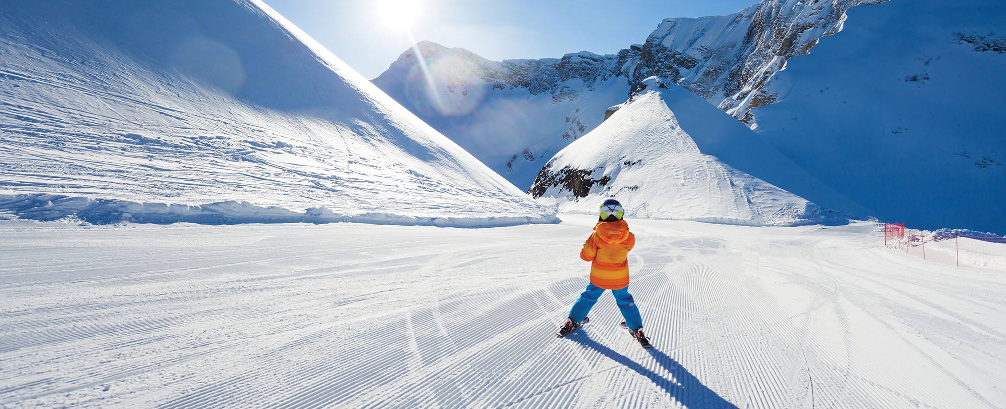 A kid in a bright orange jacket skiing down a freshly groomed ski slope on a sunny day