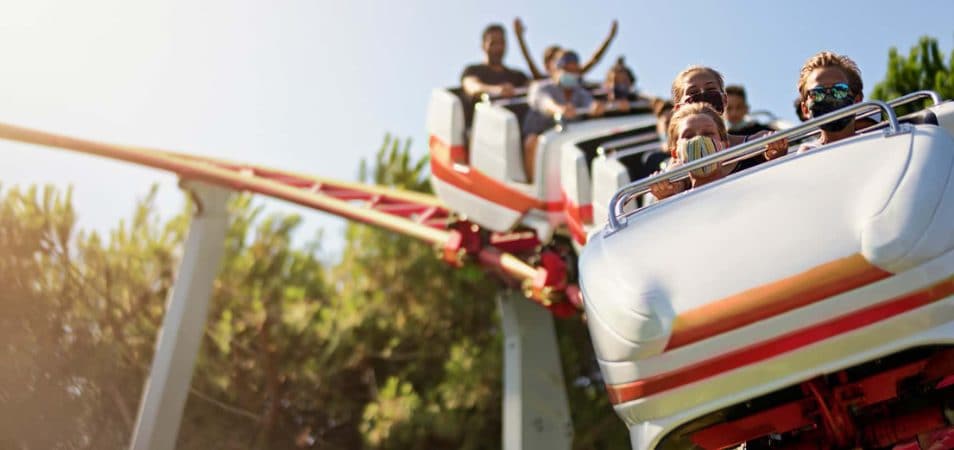 Three mask-wearing kids ride in the front car on a white and red roller coaster