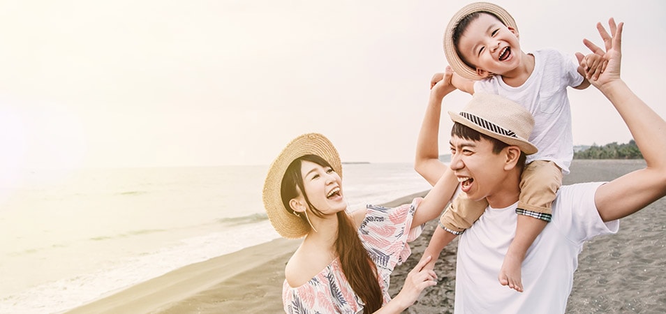 A young family laughs together on a sunny beach while dad holds their son on his shoulders