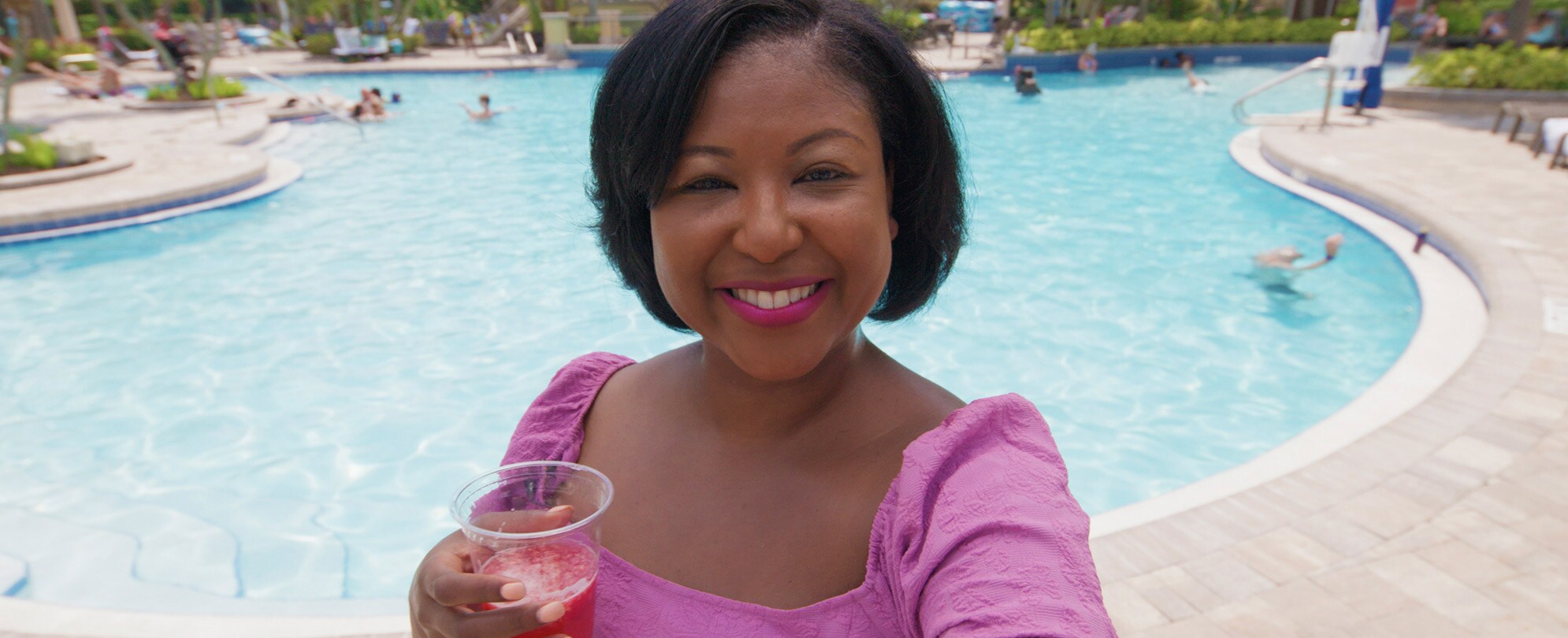 A woman is taking a selfie while drinking a cocktail in front of a pool.