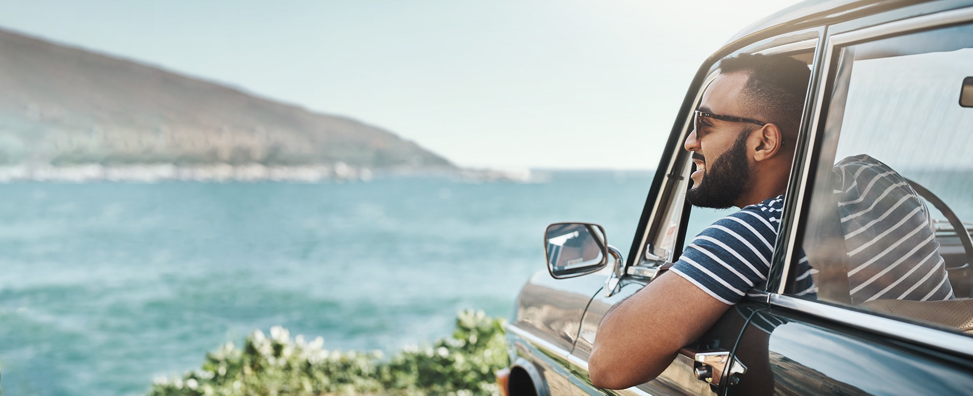 A man leans out the window of his car smiling as he looks out over the shore on a sunny day.