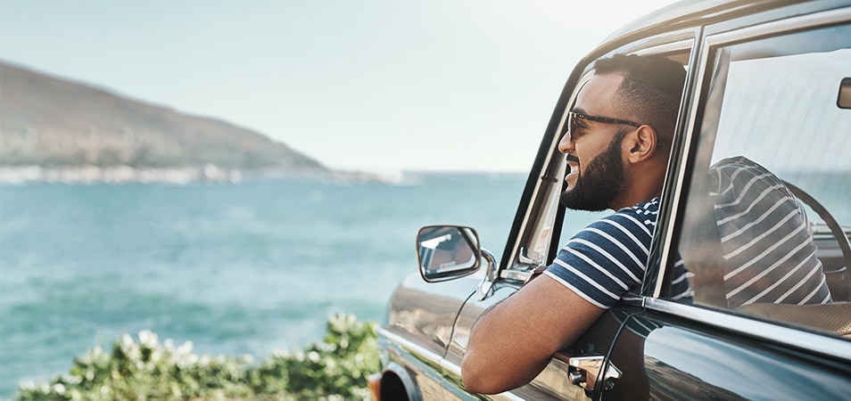 A man leans out the window of his car smiling as he looks out over the shore on a sunny day.