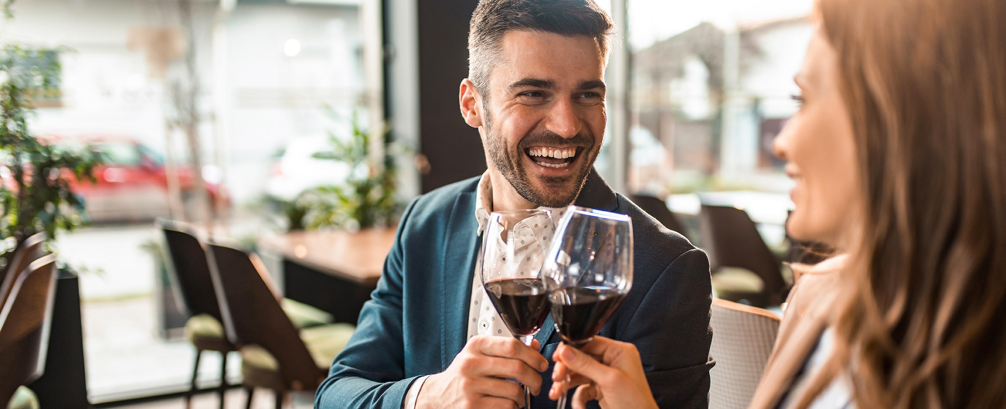 A man smiles at a woman as they clink glasses of red wine while sitting in a restaurant.