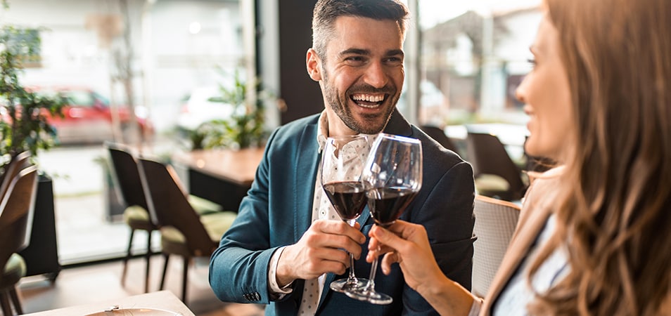 A man smiles at a woman as they clink glasses of red wine while sitting in a restaurant.