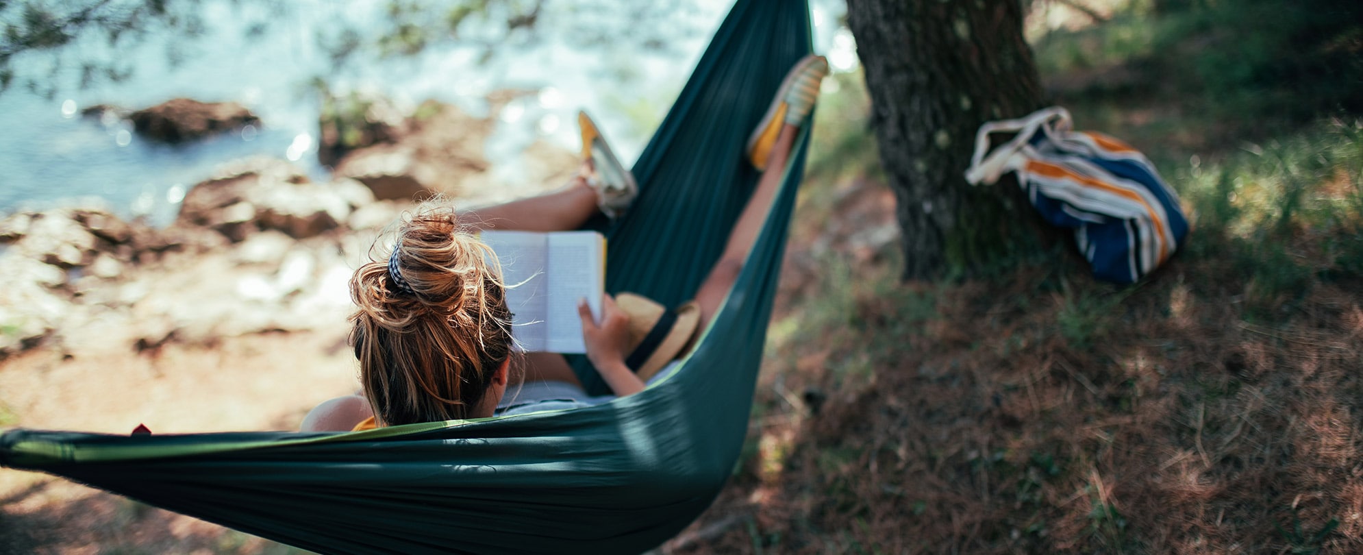 View from behind as a woman reads a book while lounging in a hammock next to a stream.