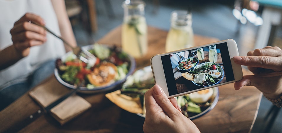 Woman holds her phone above a table for two to take a photo of their meal on a wooden table, including two full plates of food and two glass jars of lemonade.
