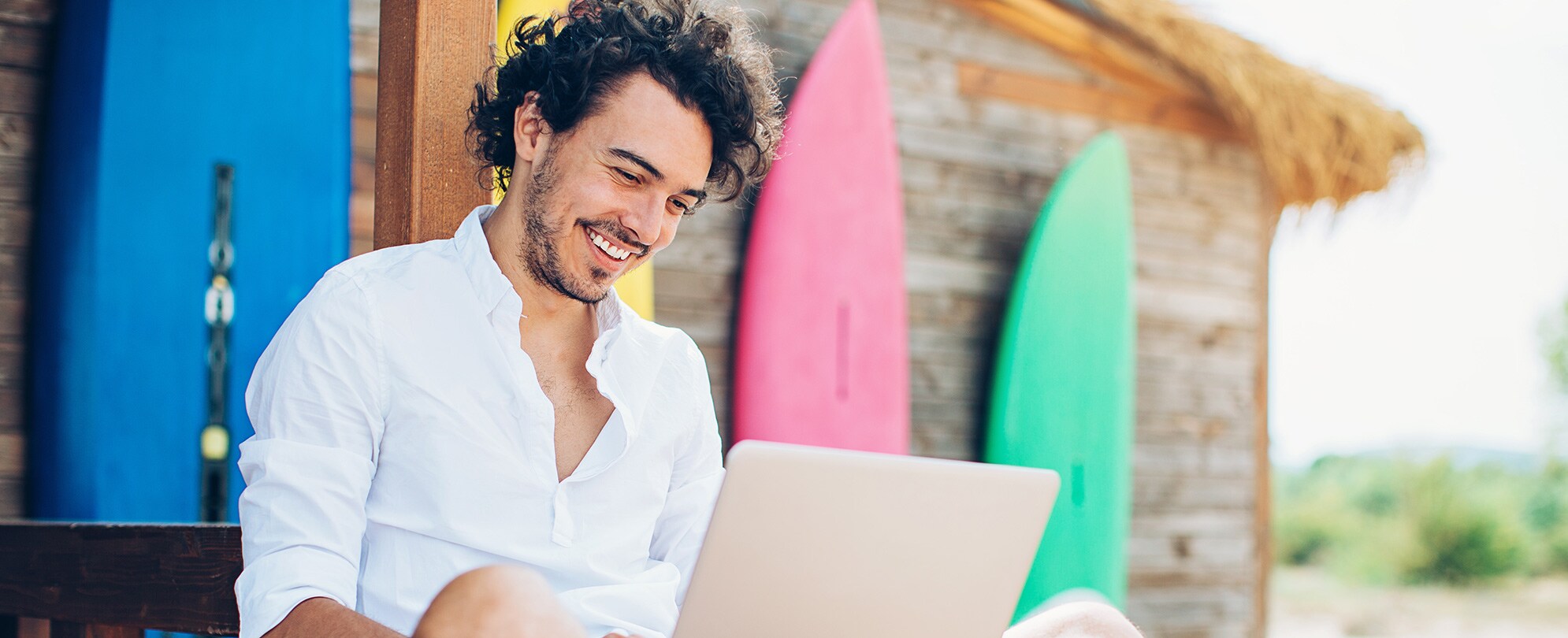 Man sitting near a surfboard shed and smiling while looking down at a laptop.