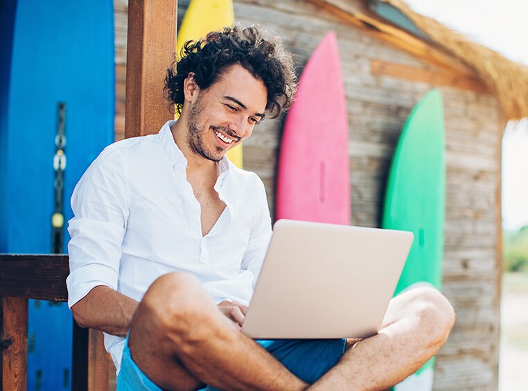 Man sitting near a surfboard shed and smiling while looking down at a laptop.
