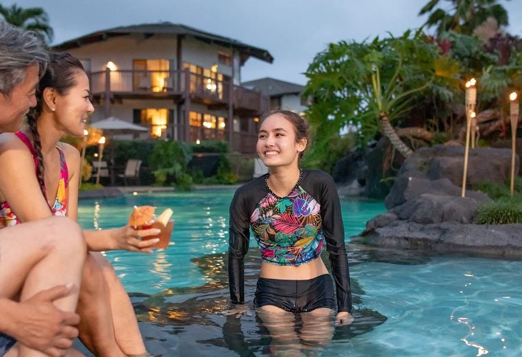 A mom and dad, sitting outside of a pool enjoying a drink and conversation with their daughter who is sitting in the pool. 