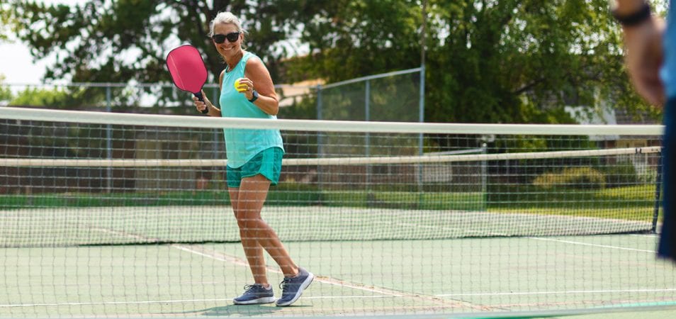 A woman smiles holding a pickleball paddle and ball while standing on a sports court about to serve to her partner