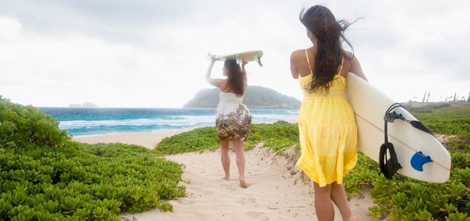 Two women surfers wearing sundresses carry surfboards down a sandy path towards the ocean in Hawaii.