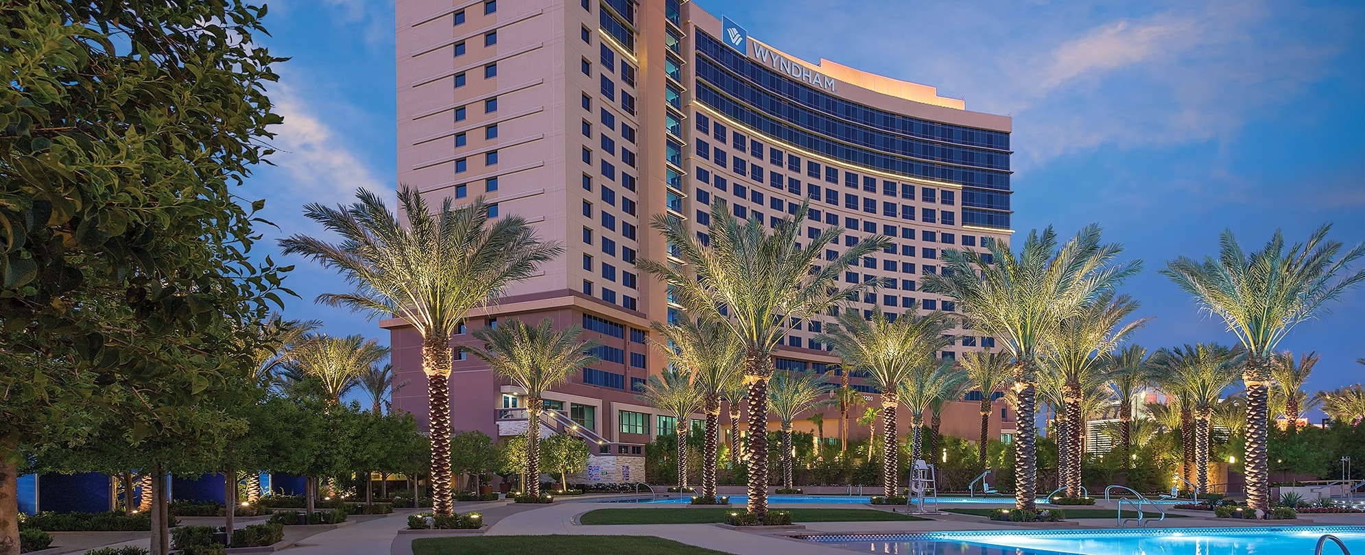 The Wyndham Desert Blue rises against an evening sky behind elegant landscaping dotted with swimming pools and up-lit palm trees