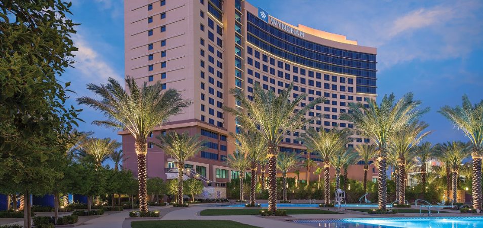 The Wyndham Desert Blue rises against an evening sky behind elegant landscaping dotted with swimming pools and up-lit palm trees