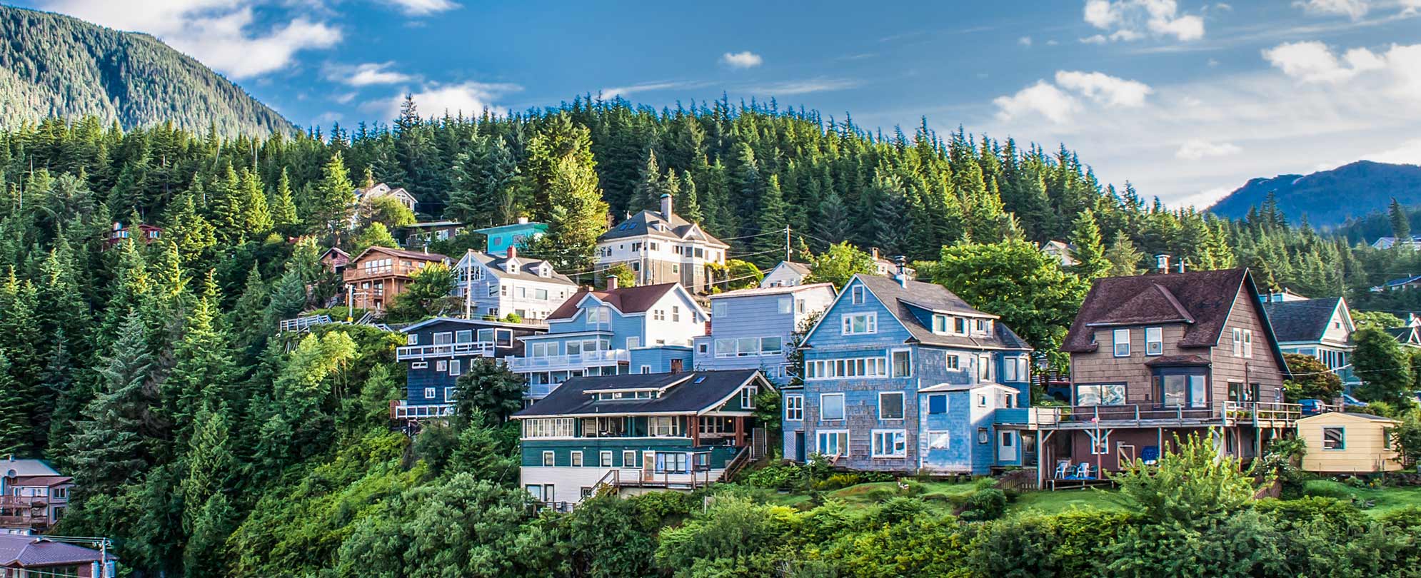 Multiple houses on a hill surrounded by pine trees and mountains in Ketchikan, Alaska.