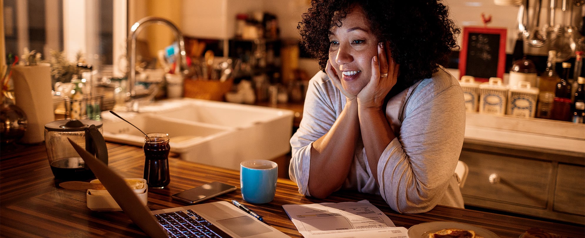 A woman smiles at her computer as she rests her elbows on the kitchen counter and holds her hands on each side of her face.
