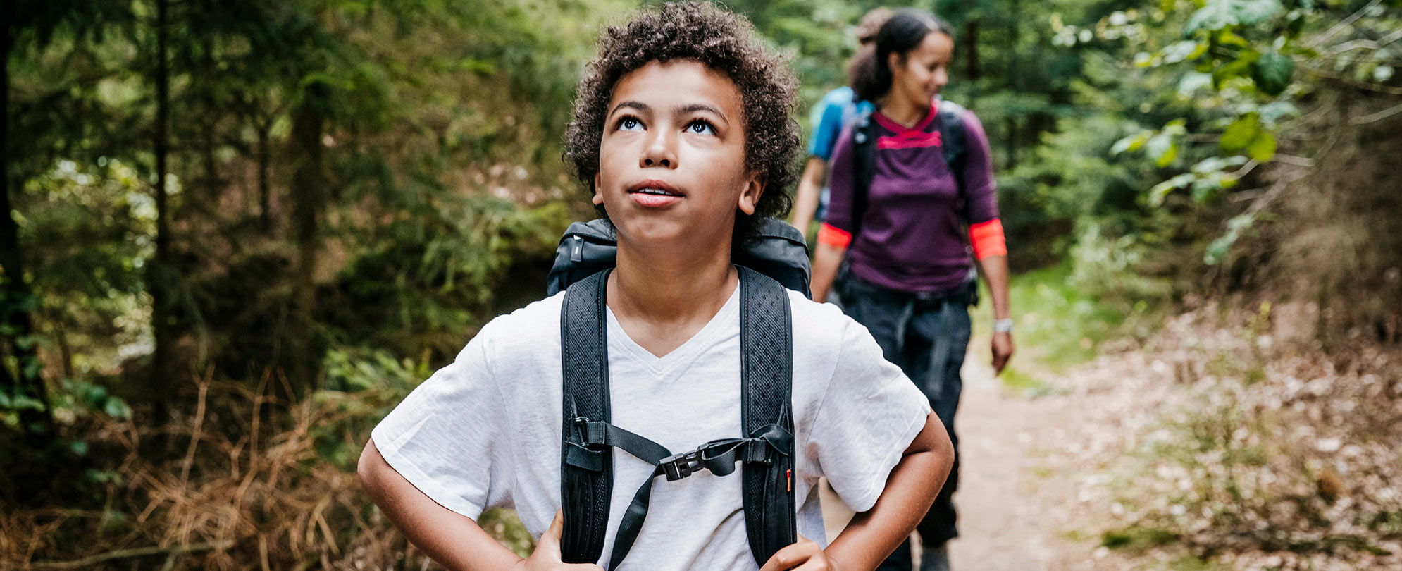 A pre-teen boy takes in the views as he backpacks on a trail through the forest with a young woman and man following behind him.
