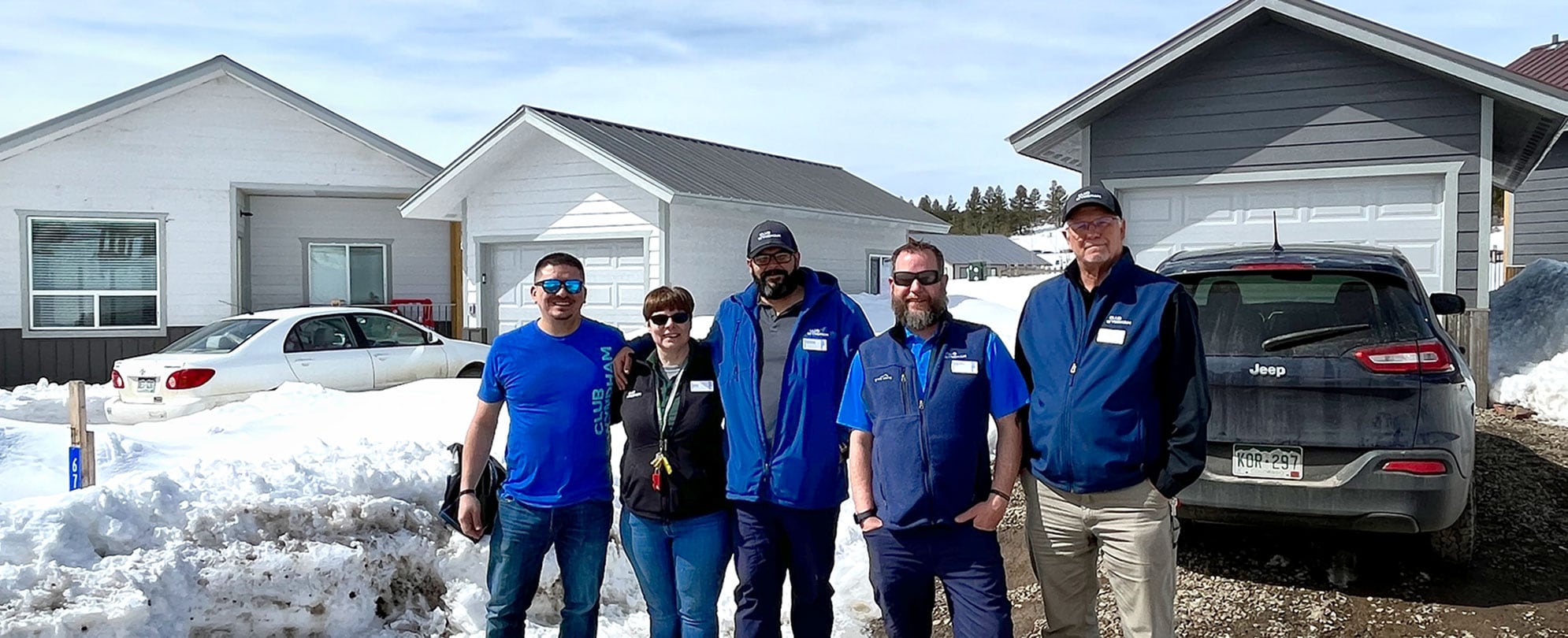 Employees from the WorldMark Pagosa resort, four men and one woman, stand in front of a pair of newly built houses with a dark car in one driveway, a white car in the other driveway, and large amounts of snow piled up in between the houses. 